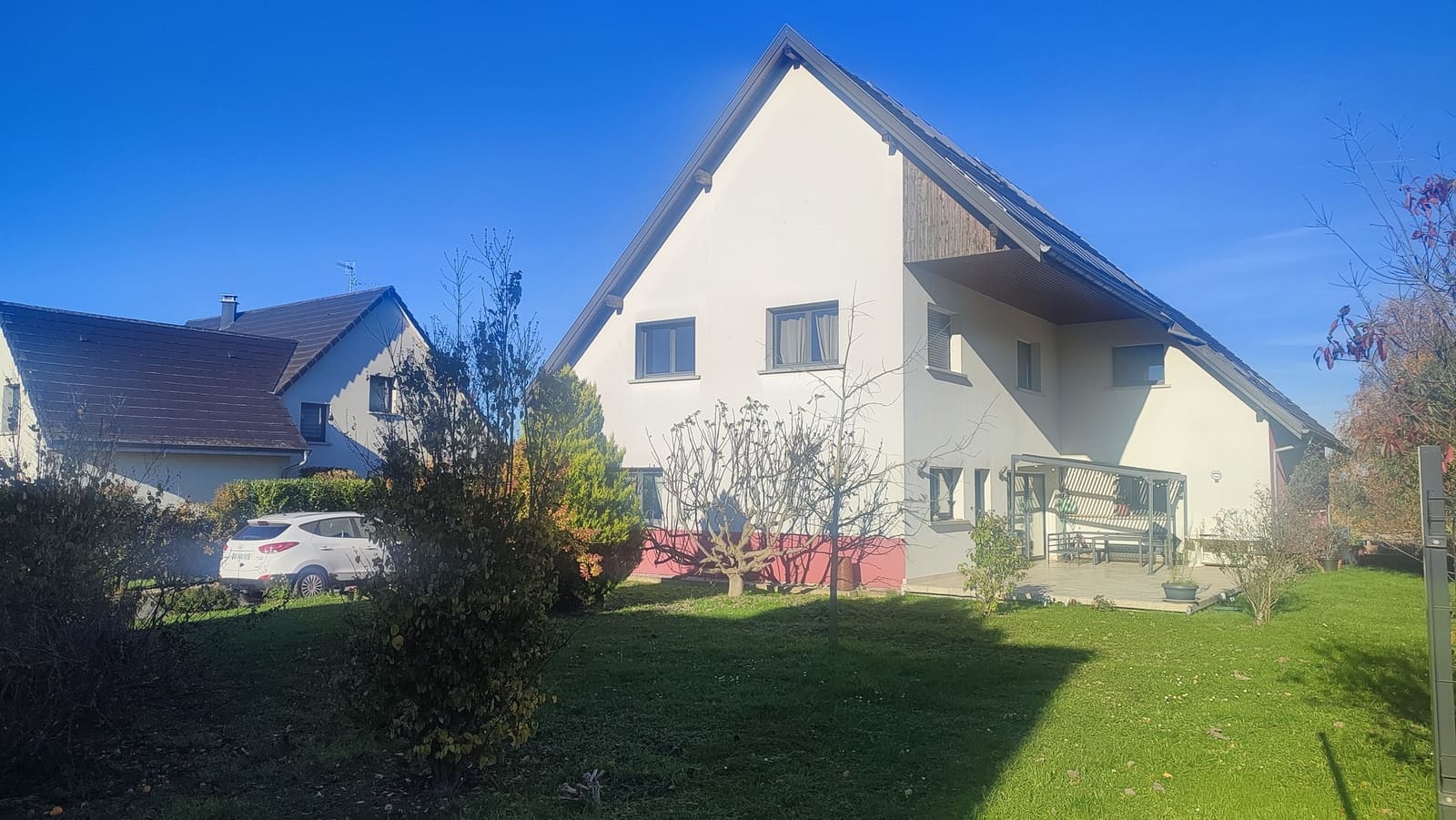 Très grande maison familiale à vendre à Saint-Bernard, en Alsace, vue en façade sur un vaste jardin arboré bien entretenu. La maison, de style contemporain, offre une architecture équilibrée avec un toit en pente, des façades claires et de grandes fenêtres qui laissent entrer beaucoup de lumière naturelle. On distingue une terrasse accueillante, un espace de stationnement et un environnement résidentiel calme, idéal pour une famille. Le terrain d’environ 2 000 m² entoure la maison et propose une belle pelouse, des arbustes et des arbres, offrant de nombreuses possibilités d’aménagement extérieur, jeux pour enfants, potager ou coin détente. Cette maison d’environ 200 m² habitables, avec sous-sol, rez-de-chaussée et étage, représente une opportunité rare dans le secteur de Saint-Bernard, à proximité de Mulhouse et de Bâle, pour un projet de vie confortable et lumineux.